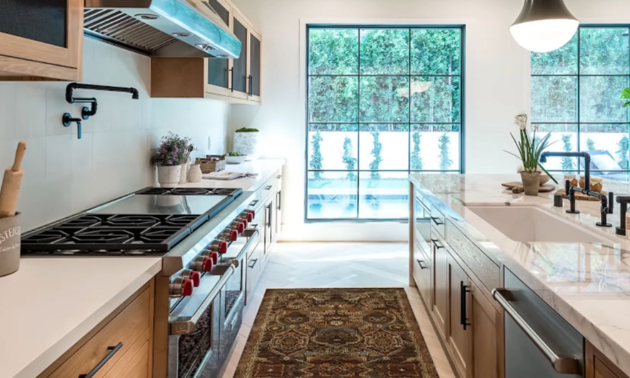 A luxurious, well-lit galley kitchen with light wood cabinets, white countertops, a large stainless steel range, and a marble island. A black-paned window overlooks lush greenery, and a patterned runner rug lies on the pale wood floor.