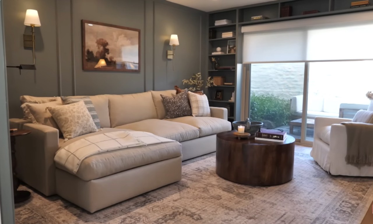 A view of a living room with a large, comfortable beige sectional sofa with a chaise lounge covered by a plaid throw blanket. The room has dark sage green walls with decorative paneling and is anchored by a patterned area rug. A round, dark wood coffee table sits in front of the sofa, displaying a candle and books. Built-in shelving with various decor items is visible on the right, next to a large sliding glass door.