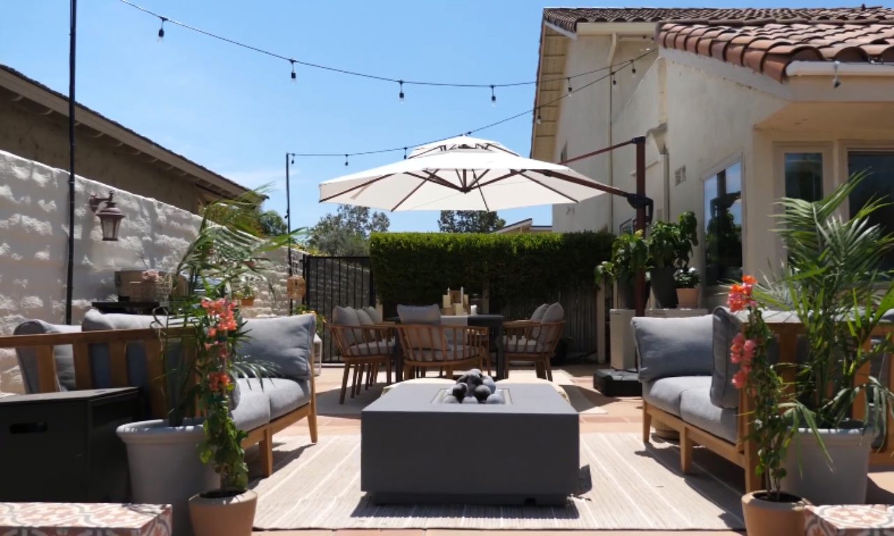 A bright, sunny outdoor patio set up for entertaining. The space features a neutral-toned rug, a dark gray square fire pit table surrounded by matching light-wood-framed lounge chairs with gray cushions, and a large white cantilever umbrella providing shade over a dining set in the background. String lights are hung across the patio, and tall planters with flowering plants define the seating area.