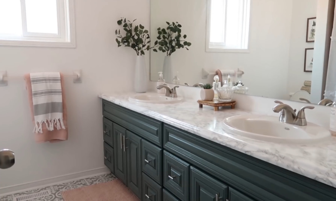 A renovated, bright bathroom featuring a double vanity with a dark teal-green cabinet base and a white, marble-patterned countertop with two sinks. A large mirror reflects the opposite wall with two bright windows. A vase with faux eucalyptus branches sits on the counter. The new, patterned floor tiles are visible at the bottom.