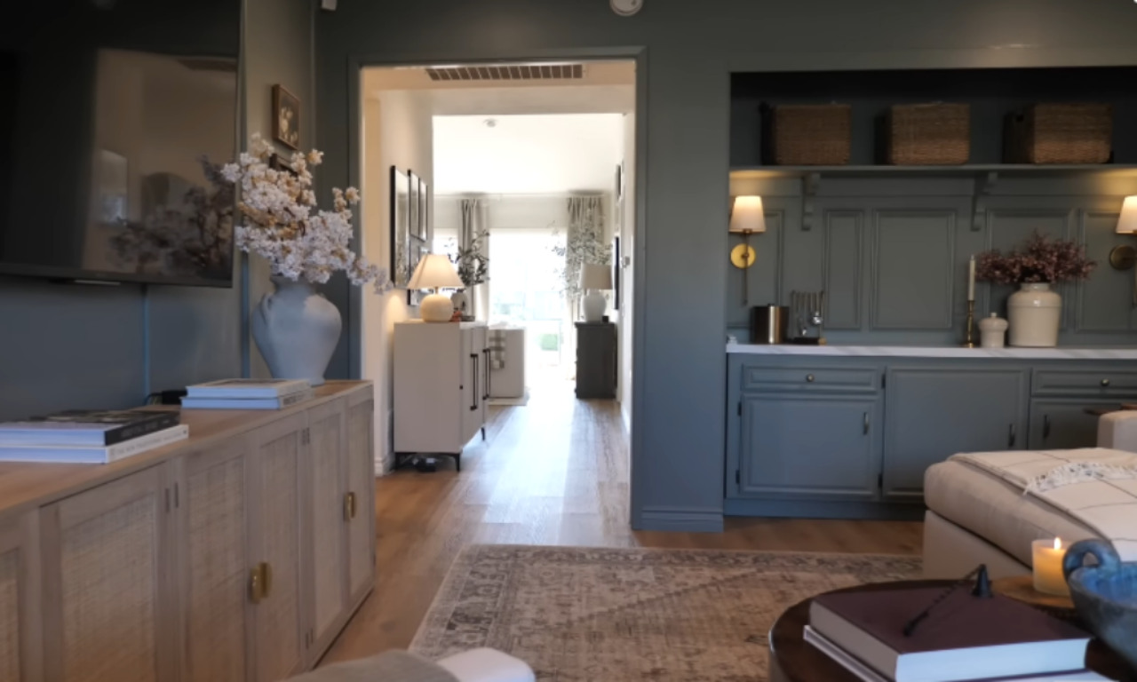 An interior view of a living space with a focus on a recessed built-in cabinet area on the right and a hallway leading into another room. The walls are painted a deep gray-green color. The built-in cabinet area features lower closed storage painted the same color with a white counter and upper shelves holding woven baskets, illuminated by two gold wall sconces. A light wood media console with a large white floor vase is on the left, and a patterned area rug partially covers the wood floor.
