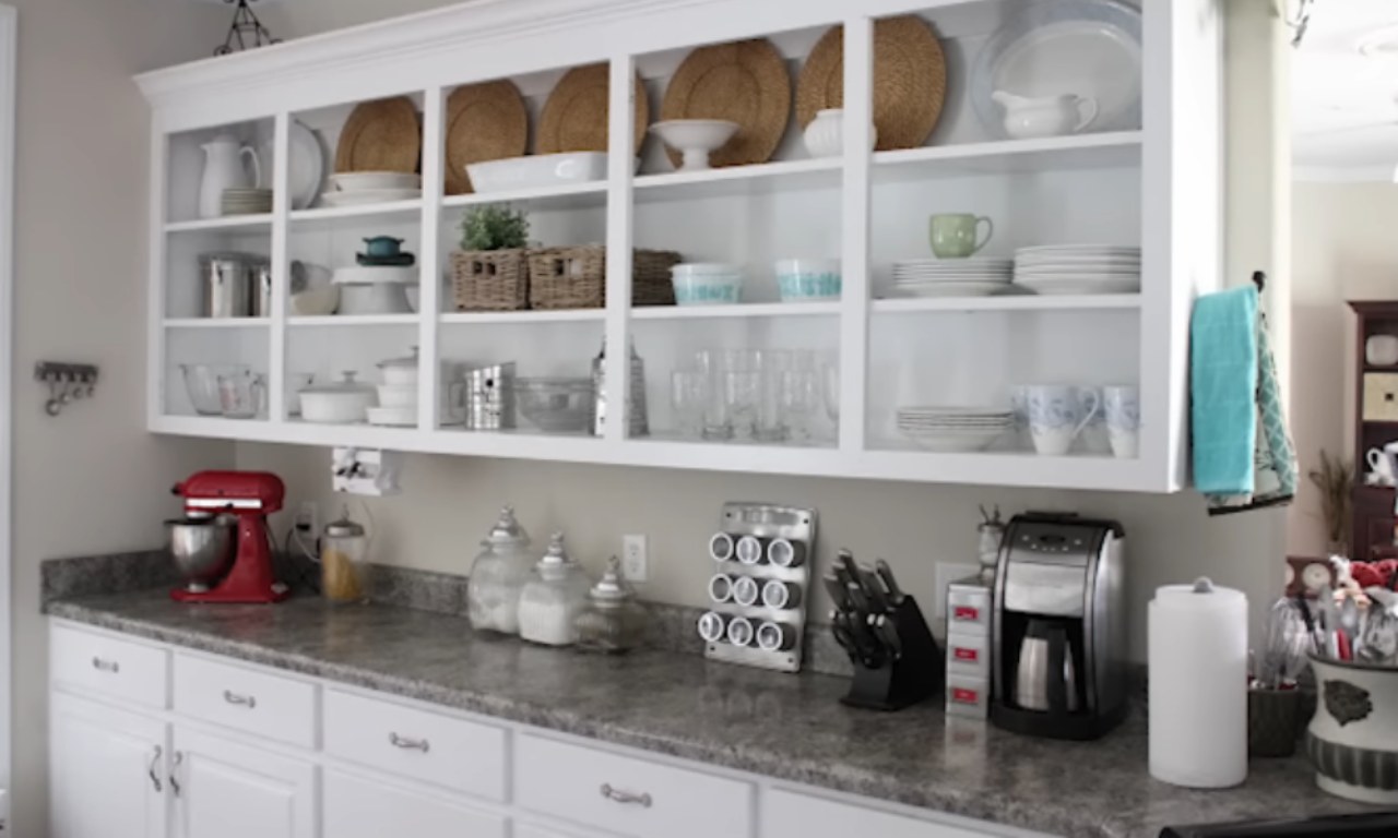 A bright kitchen with white cabinets. The upper cabinets are open shelving, displaying various dishes, plates, and small decorative items. The lower cabinets have a grey countertop with a red stand mixer and a coffee maker. The scene illustrates renter-friendly decorating ideas.