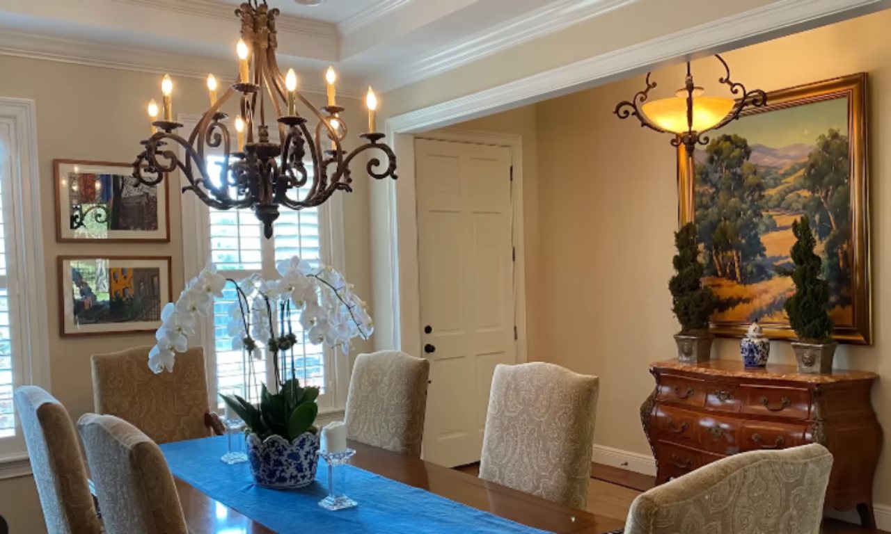 A traditional dining room with beige walls and a wooden dining table set for a meal. A large, ornate wrought-iron chandelier hangs over the table. The table is dressed with a blue runner and a centerpiece of white orchids in a blue and white planter. In the background, there is a large framed landscape painting and a dark wood buffet.