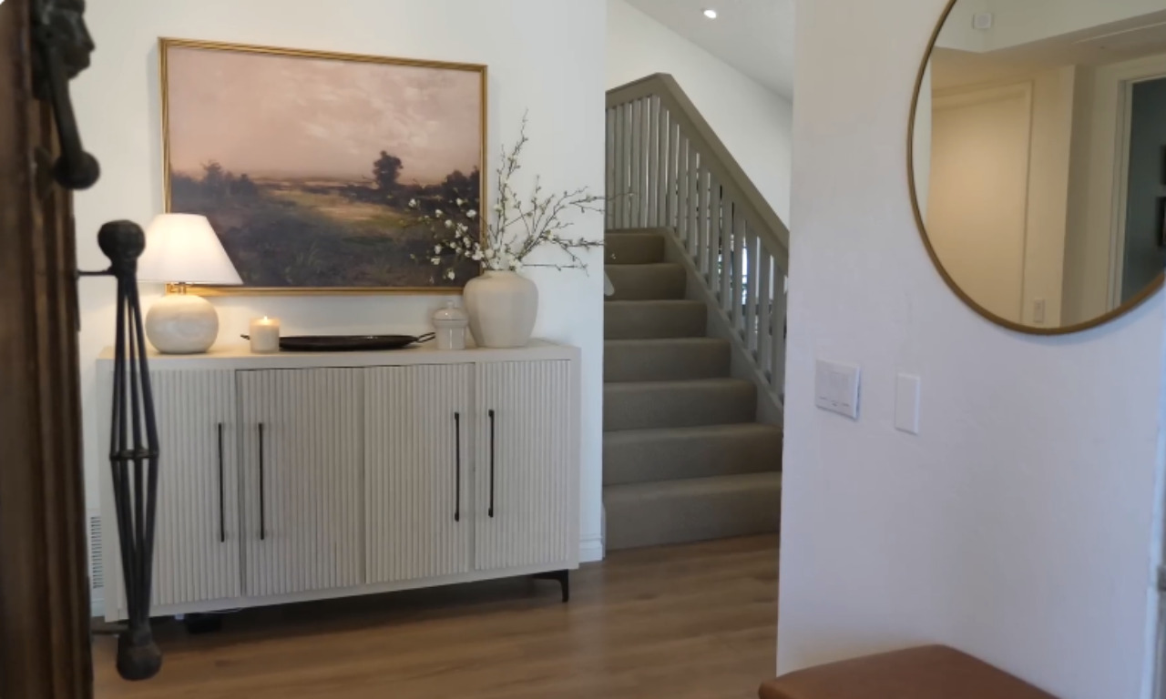 A modern entryway features a textured white console cabinet with black hardware, topped with a landscape painting, a white lamp, a vase with white flowers, and a candle. A carpeted staircase is visible in the background, and a round mirror is on the wall to the right.