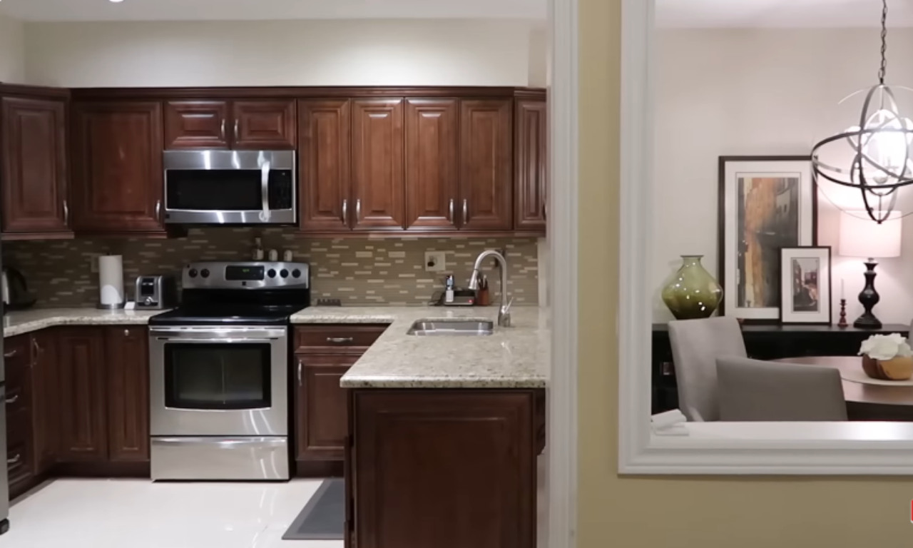 An interior view showing a brown wooden kitchen with stainless steel appliances (microwave, oven/range, refrigerator, toaster) and light speckled granite countertops. The kitchen has a mosaic tile backsplash. In the foreground, there is a cutout opening in the wall, revealing a glimpse of a decorated room with a dining table, chairs, and a large decorative vase.