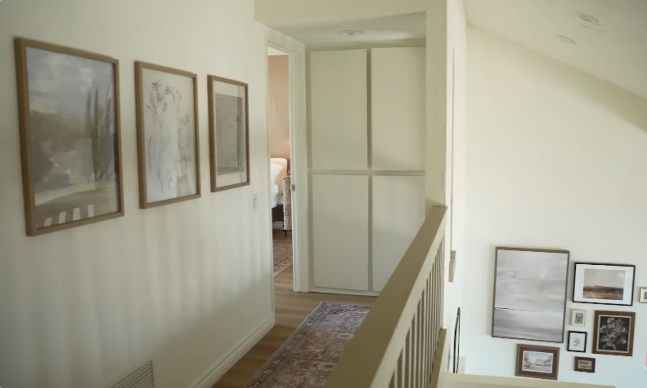 A well-lit interior hallway and staircase landing area. The hallway features three framed, nature-themed art pieces hanging vertically, a runner rug, and leads to a closet with white paneled doors. The staircase landing below has a large gallery wall of various framed artworks.