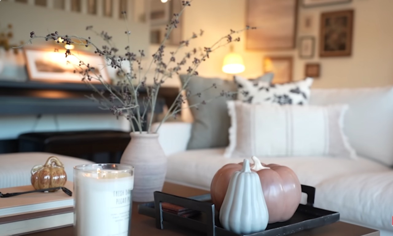 A close-up view of a cozy coffee table centerpiece. It features a lit candle, a small decorative pumpkin on a stack of books, and a vase holding dark-colored berry branches. In the background, there is a black tray with two ceramic pumpkins (one pale pink and one white/light blue) and a white sofa with pillows.