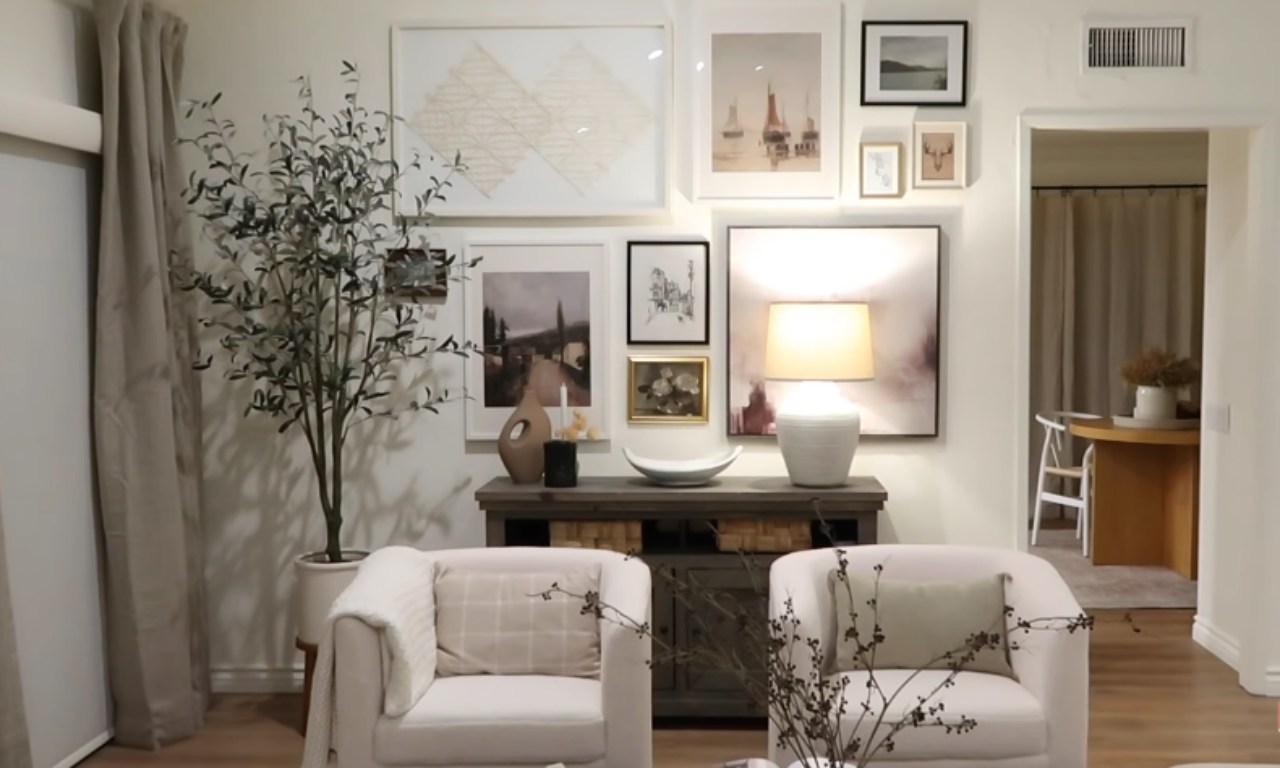A cozy living room area featuring a large, eclectic gallery wall hung above a dark wooden console table. Two light-colored, upholstered armchairs sit in front of the console. A tall, faux olive tree stands to the left. The color palette is neutral, with shades of white, beige, and brown.