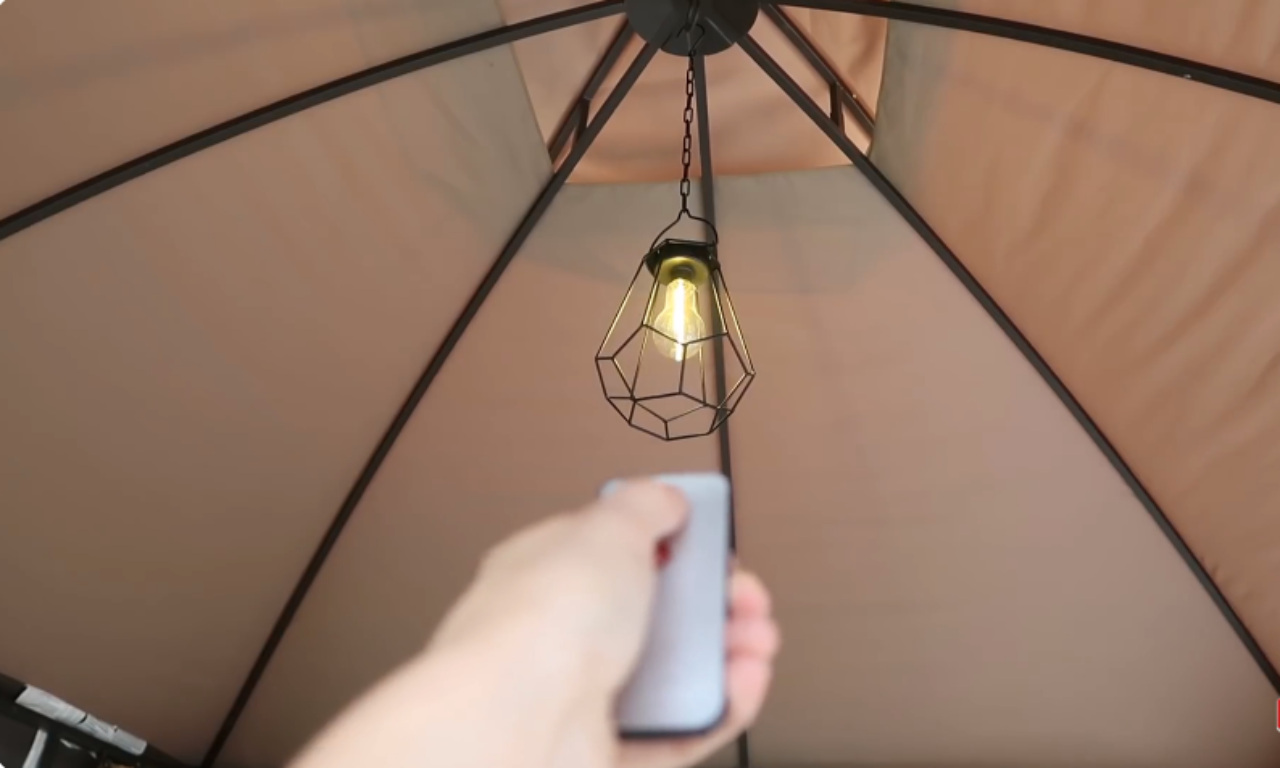 A hand holding a remote control, pointing up toward a geometric pendant light with an exposed Edison bulb hanging inside a gazebo.