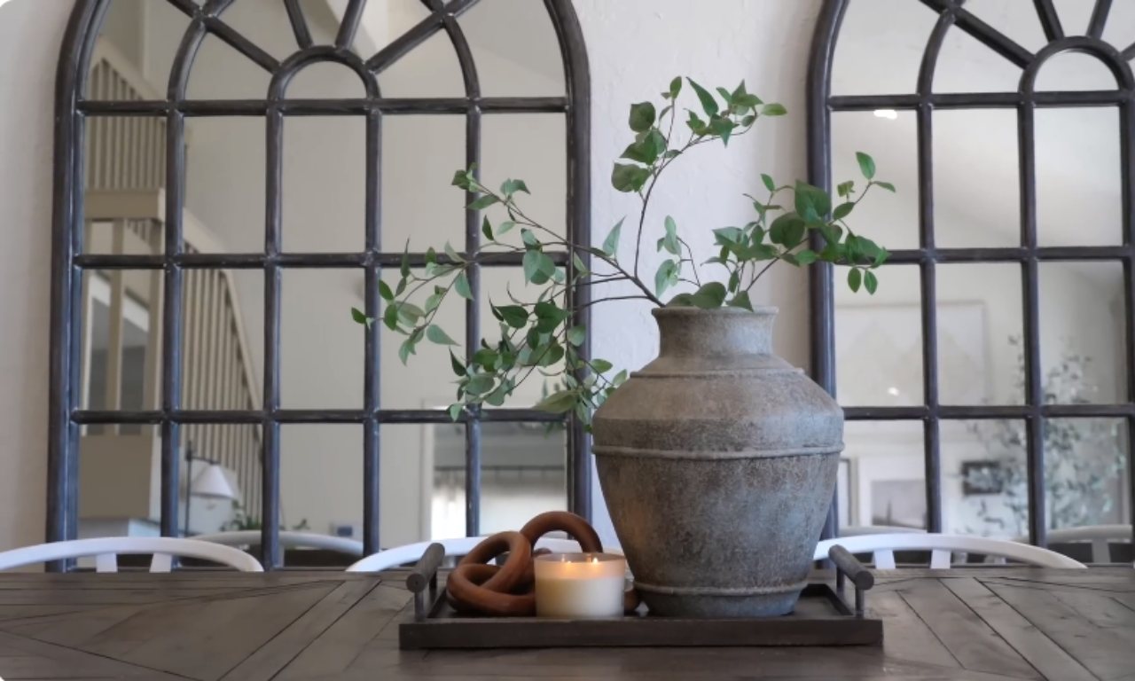 A rustic, galvanized vase with greenery sits on a dark wooden tray on a dining table, next to a lit candle and a decorative wooden knot. In the background are two large, arched, multi-paned wall mirrors.