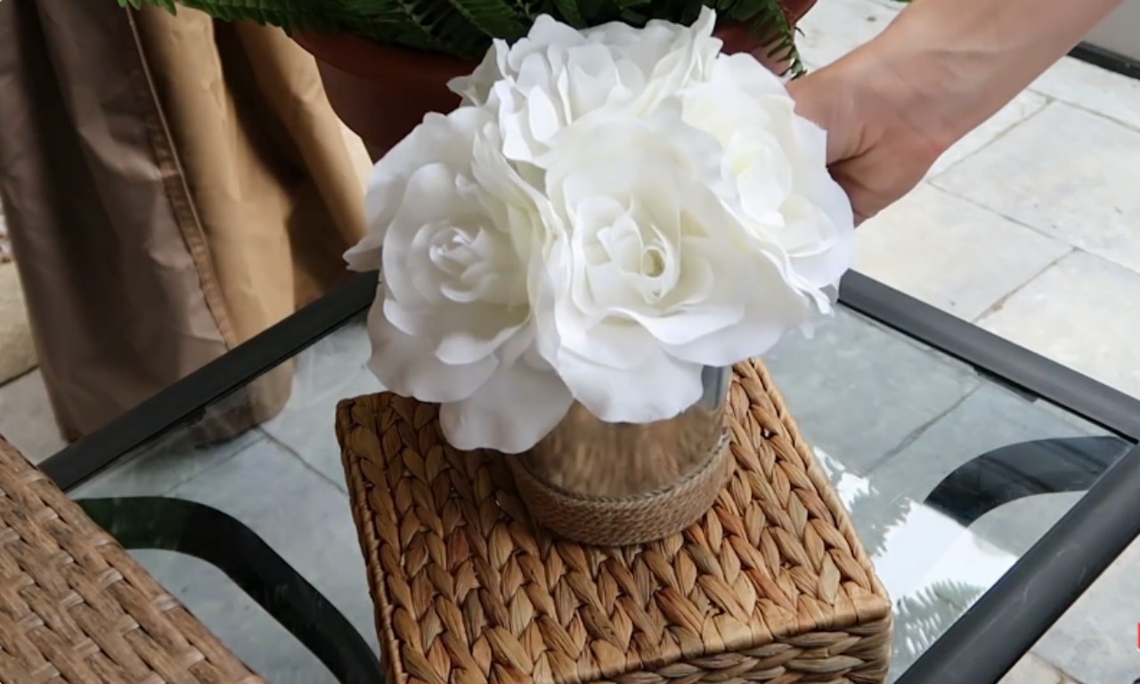 A close-up of a hand placing a glass jar filled with white artificial roses on a woven wicker box on an outdoor table.