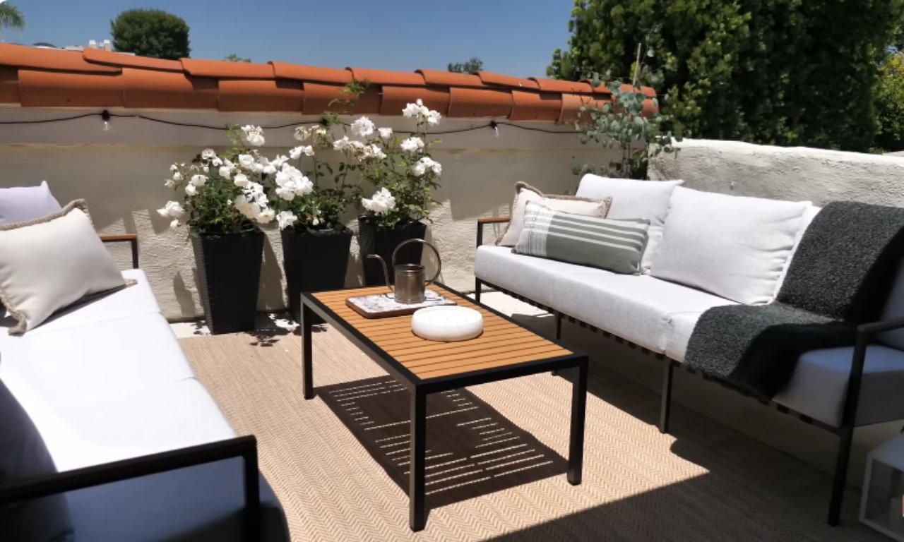 A bright outdoor patio space furnished with a gray sectional sofa and a matching outdoor chair, set upon a beige woven rug. The patio features a dark wood and black metal coffee table, and tall black planters with white flowers and greenery against a low, white stucco wall topped with terra cotta roof tiles.