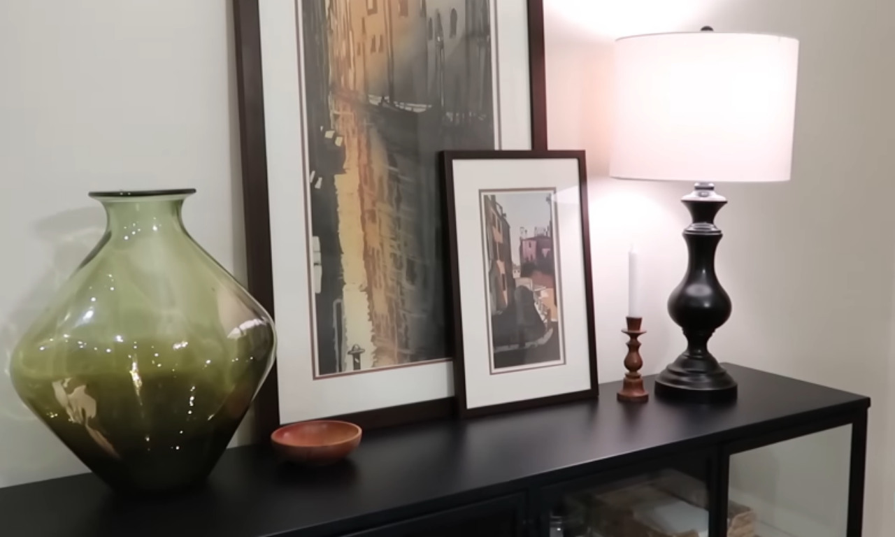 A close-up view of a dark wooden console table decorated with three main items: a large, rounded, translucent olive green glass vase on the left, a tall black table lamp with a white cylindrical shade on the right, and two framed watercolor-style paintings leaning against the wall. A small wooden bowl and a slender white candle in a wooden holder sit between the items.