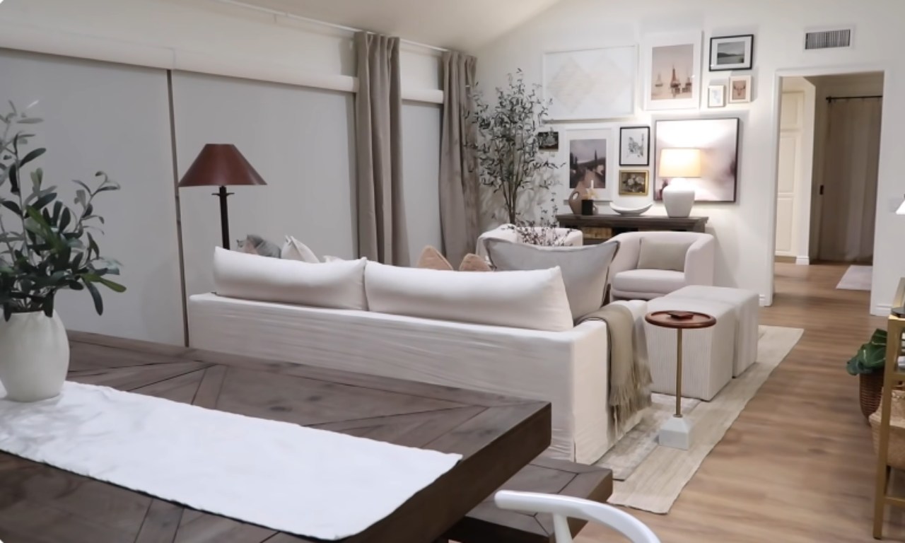A wide view of a modern living room and dining area, demonstrating layered lighting and a neutral color scheme. A large, white slipcovered sofa is centered in the living area, facing a dark wooden dining table in the foreground. Heavy beige drapes frame the window wall. The gallery wall and two armchairs are visible in the background.