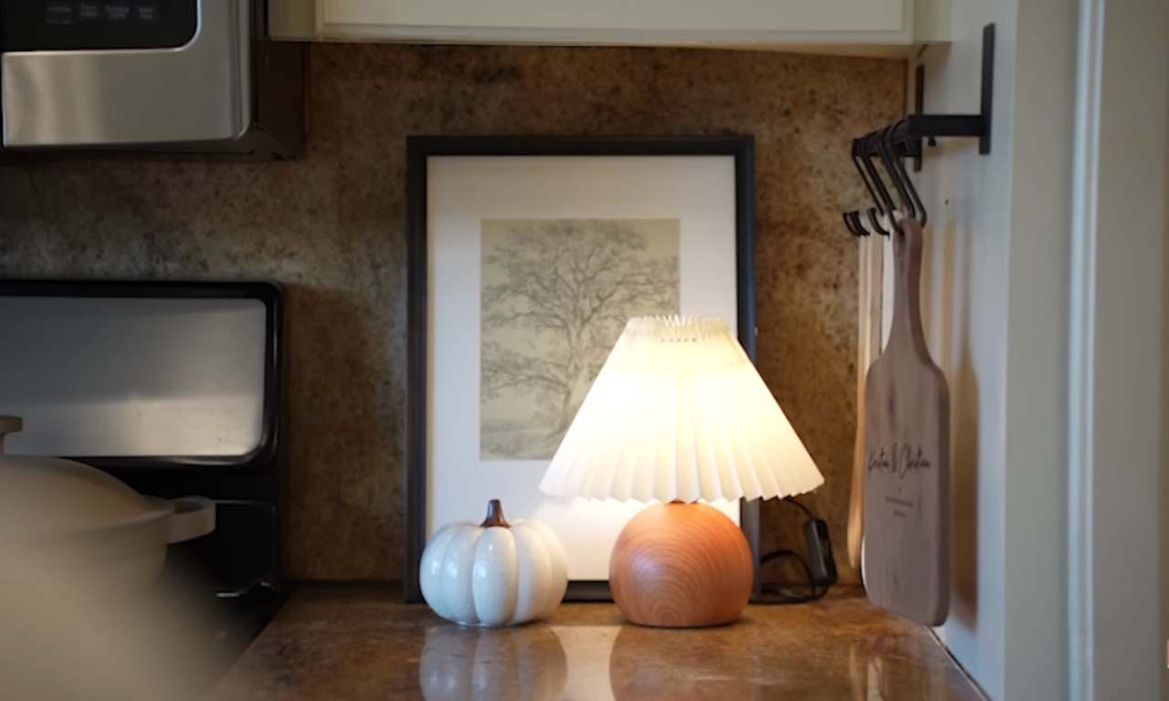 A cozy vignette in a kitchen. It shows a small wooden table lamp with a pleated white shade and a light blue ceramic pumpkin decoration sitting on a countertop. Behind them is a framed print of a tree, and to the right, a wooden cutting board hangs on the wall. The backsplash is a textured brown material.