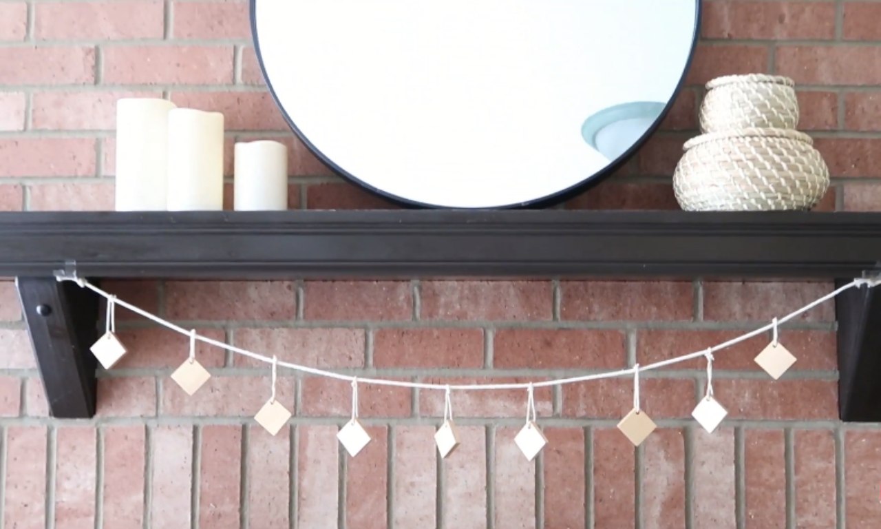 A simple, diamond-shaped wooden garland hanging across a dark wood fireplace mantel, which is set against a red brick wall. On the mantel are three white candles and two stacked woven baskets, with a large circular mirror above.