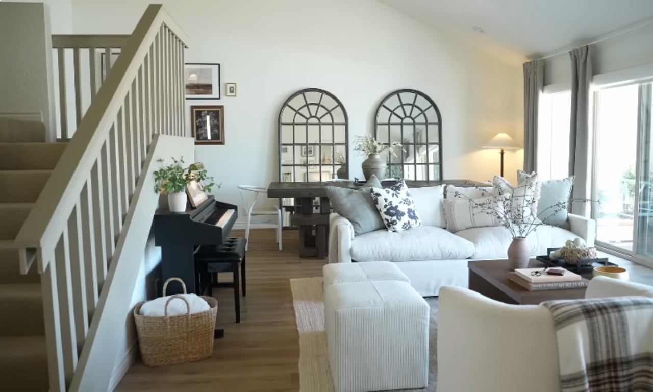 A bright, open-plan living room featuring a white slipcovered sofa and ottoman, an upholstered white accent chair, and a dining table visible behind the sofa. Two large, dark-framed arched mirrors hang on the white wall. A black baby grand piano with sheet music is positioned near the staircase, which has a light wood railing.