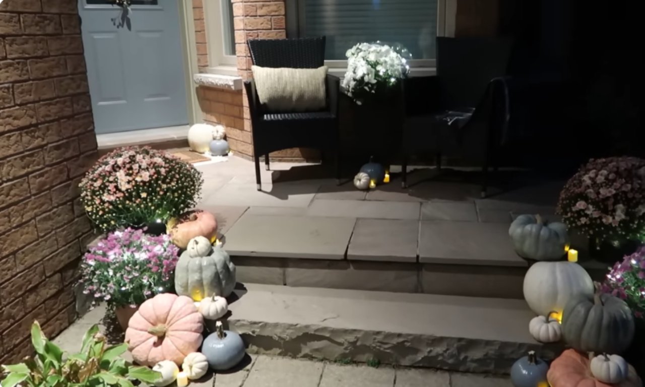 A night-time view of a front porch decorated for fall. The setting includes black wicker furniture with pillows on a raised stone platform. The lower patio and steps are heavily decorated with clusters of pastel pumpkins (pinks, blues, and whites) and large potted chrysanthemum plants, all illuminated by a soft, magical glow from scattered small candles or tea lights.