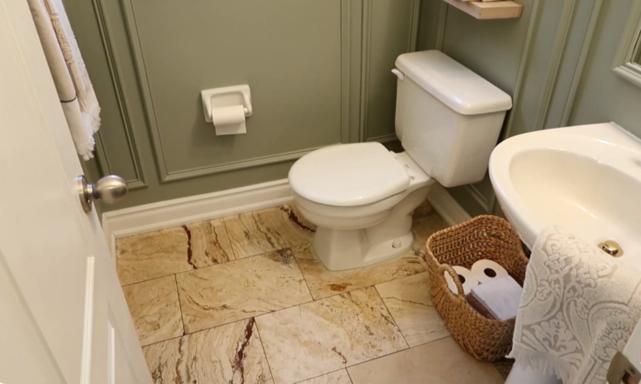 A view of the floor and lower portion of a small bathroom. The floor is covered in large, light beige and brown marbled tiles. A white toilet sits against the sage green paneled wall. A toilet paper dispenser is mounted on the wall above the toilet. A woven basket sits next to the toilet and a small white sink is visible on the right.