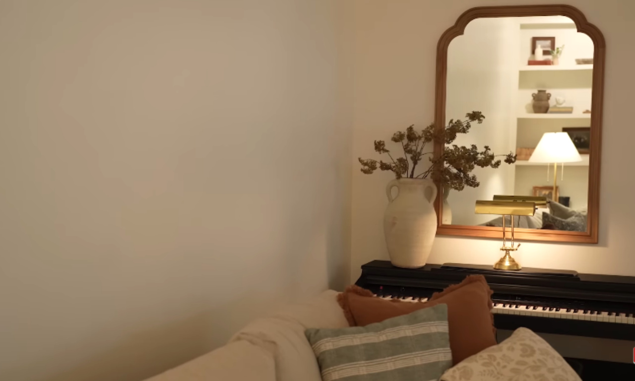 A close-up of a cozy corner with a black piano topped with a tall, light-colored ceramic vase holding dried branches. The piano is positioned next to a cream-colored couch with various throw pillows. Above the piano is a large, arched wooden mirror reflecting a softly lit built-in bookshelf area.
