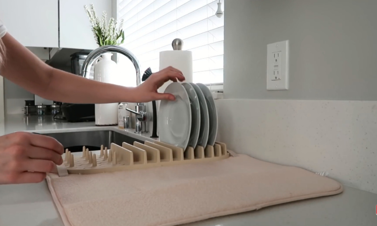 A close-up of hands placing a white plate onto a neutral-toned, slatted plate drying rack that is sitting on a beige drying mat next to a kitchen sink and faucet.