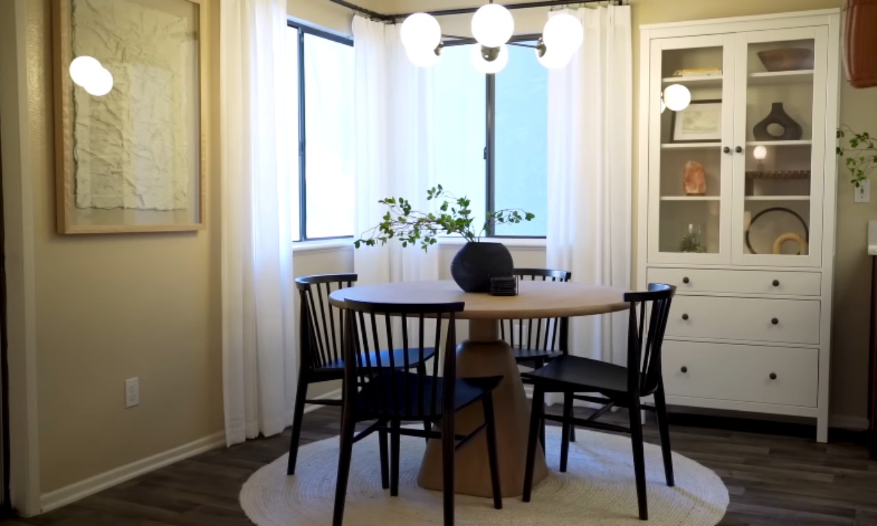 A dining area setup with a round light wood table on a circular woven rug. Four black wooden dining chairs are tucked in. Behind the table is a tall, white display cabinet with glass doors. The area is illuminated by a modern globe chandelier and natural light coming through sheer white curtains in a bay window.