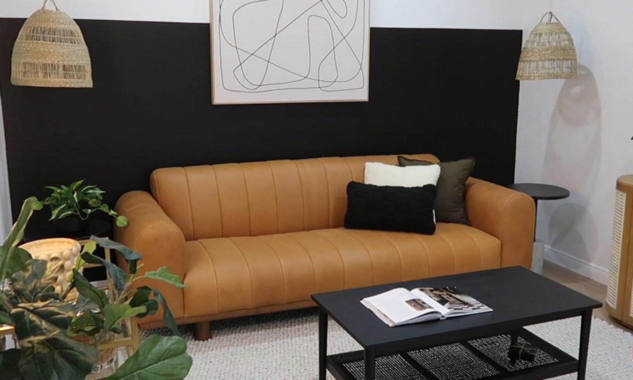 A close-up view of a living room with a dramatic black accent wall, featuring a camel-colored leather channel-tufted sofa, a black coffee table, and two woven pendant lights. The wall above the black paint is white, and the room has a textured white rug.