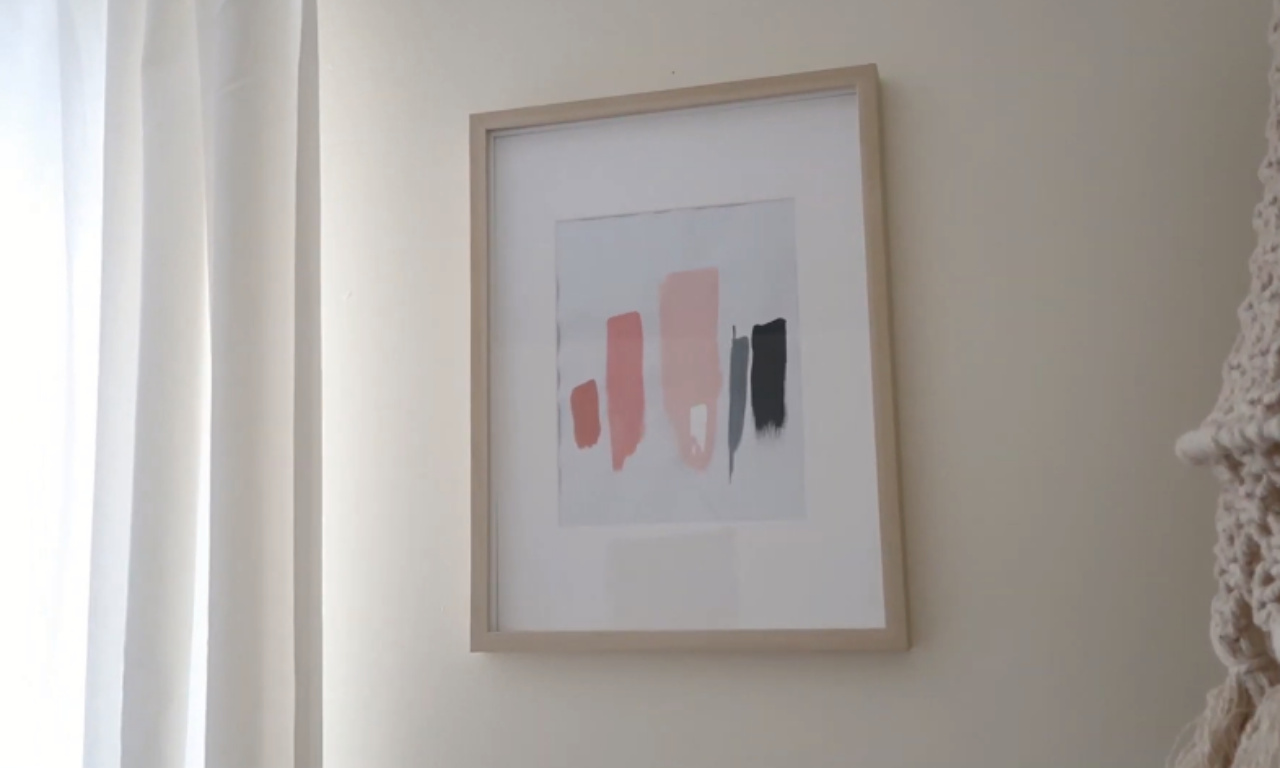 A framed piece of modern abstract art hanging on a pale wall. The artwork features vertical brushstrokes in shades of dusty rose, light pink, and black. A light-colored curtain is visible on the left.