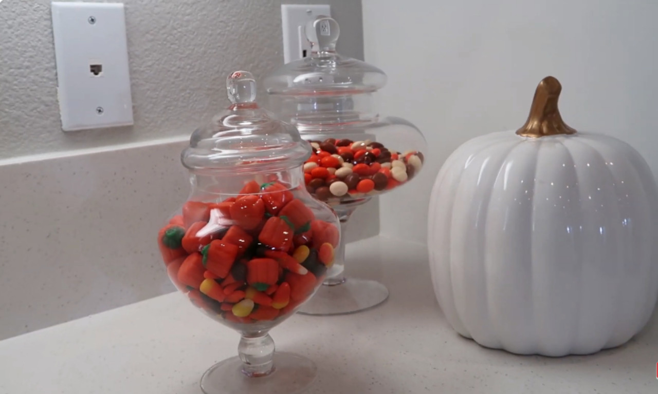 A close-up of fall-themed kitchen countertop decor. Two clear glass apothecary jars filled with colorful candies (including candy corn) are next to a smooth, white ceramic pumpkin decoration with a gold stem. The background shows a light gray wall with a white electrical outlet cover.