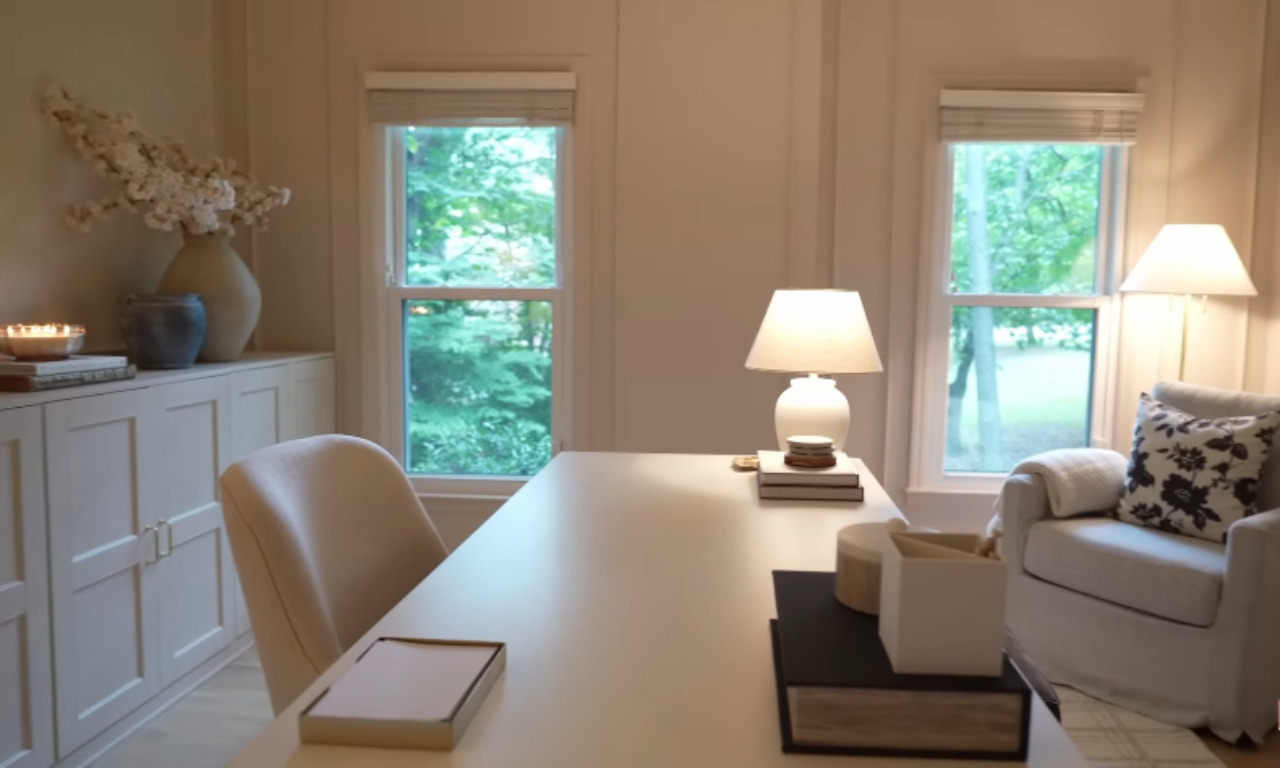 A bright, serene home office with light cream walls and white built-in cabinets on the left. A large, light-colored desk is in the center, and a comfortable armchair with a floral pillow sits by the window on the right. A white ceramic lamp lights the desk.