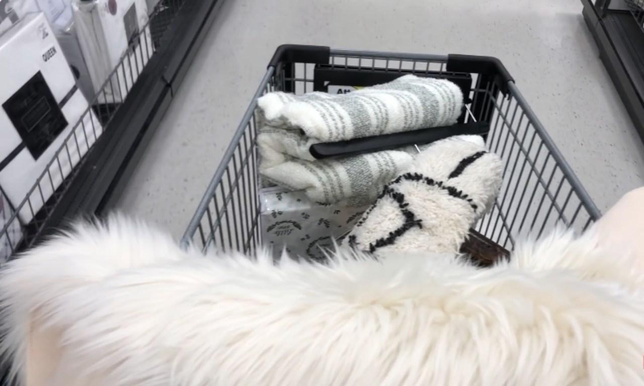 A close-up, first-person view looking down into a shopping cart in an aisle, which is filled with home decor items. Visible items include a fluffy, white faux fur blanket or rug draped over the front, and inside the cart are a folded gray and white textured blanket or throw, and a small black and white textured pillow or rug.