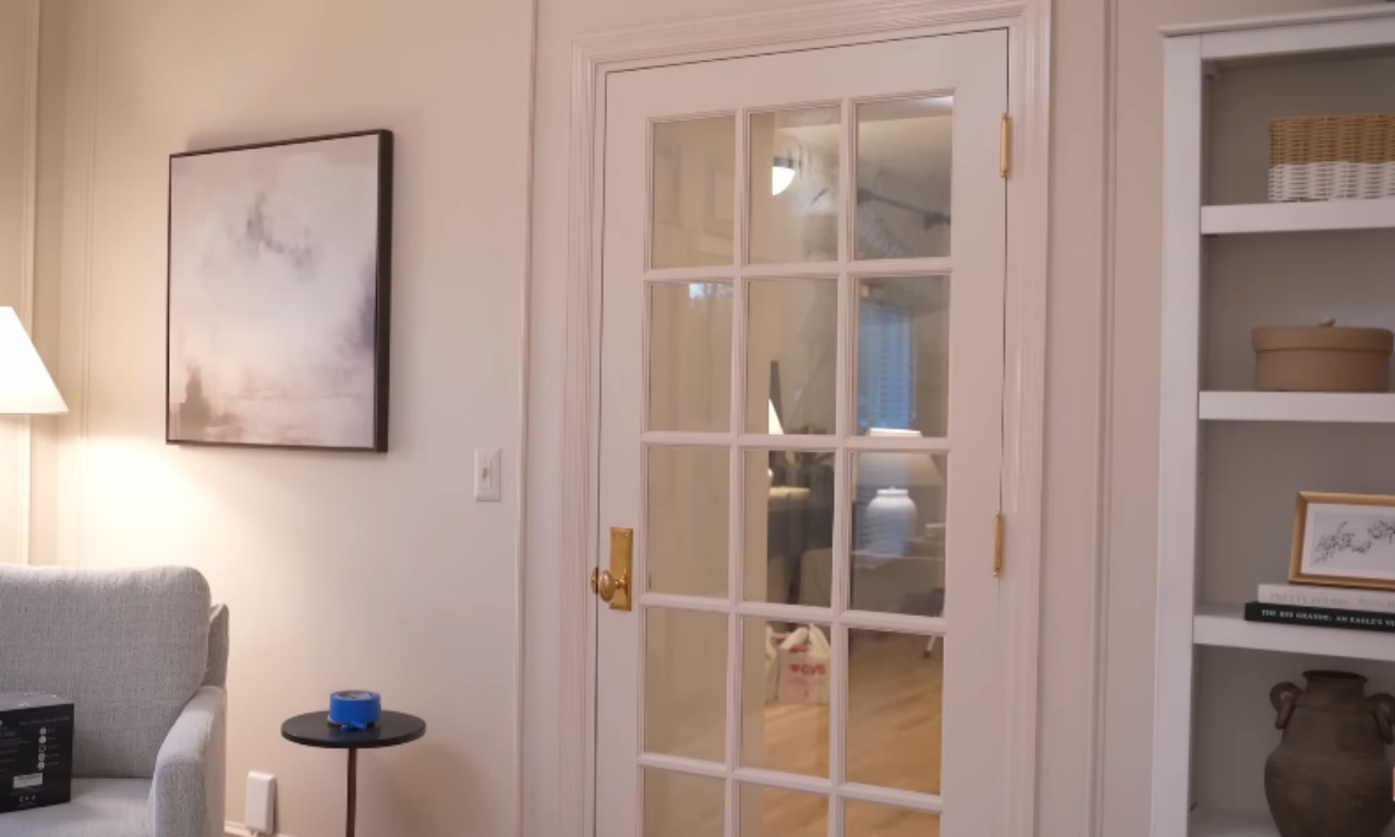 A close-up view of an interior room showing a white French door with glass panes and a gold-toned handle. To the left is a neutral-toned artwork, and to the right is a white built-in bookshelf with decorative items.