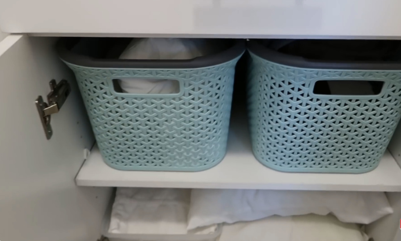 Two light blue, woven-style plastic storage baskets with gray rims sit side-by-side on a white shelf inside a white cabinet. The baskets are holding white laundry or linens, and beneath the shelf, more white linens are stored.