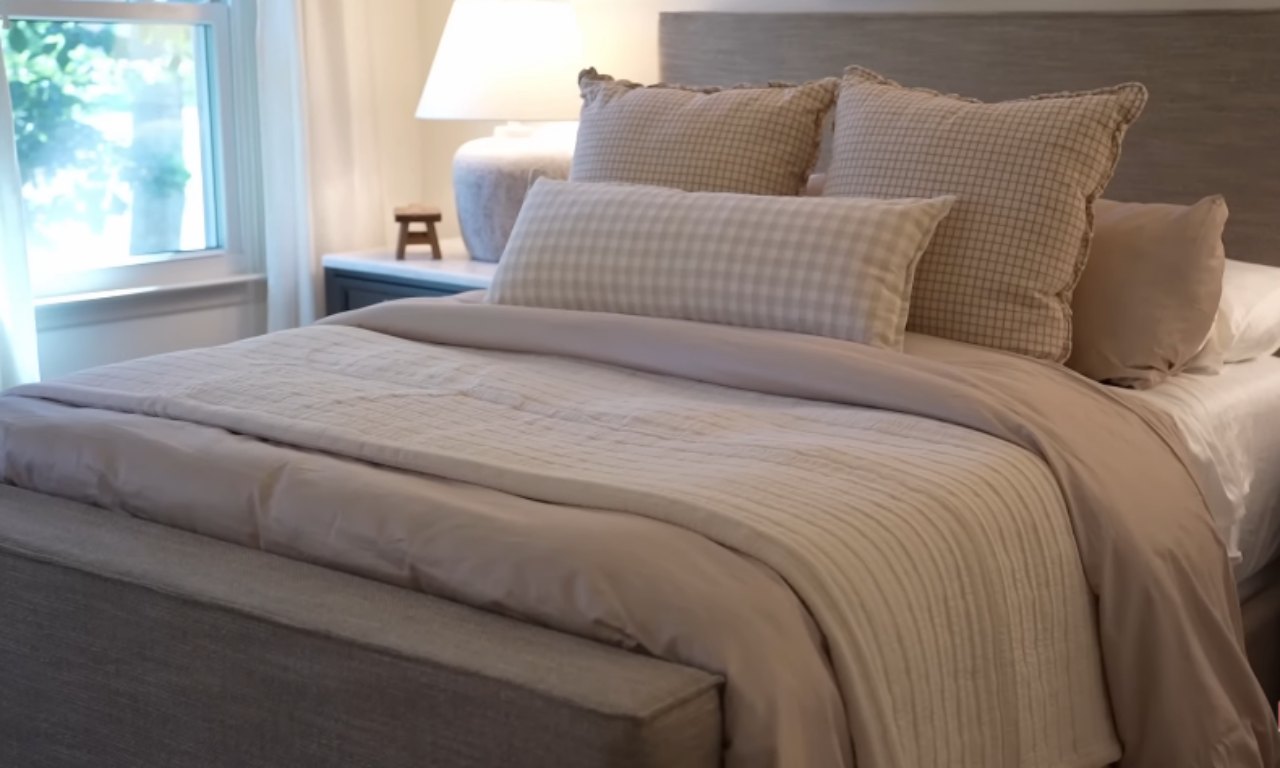 A cozy bedroom with a wooden headboard, light beige bedding, and several pillows, including one with a subtle gingham pattern. A window is visible on the left.