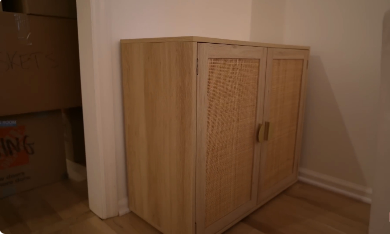 A close-up shot of a light wood cabinet with two doors featuring woven cane panels and gold handles. The cabinet is positioned against a light wall near a doorway. Cardboard moving boxes are stacked on the floor to the left of the cabinet.