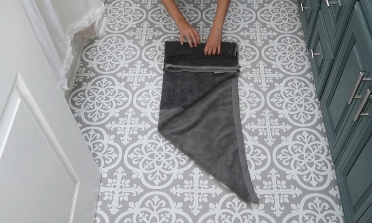 A pair of hands demonstrating a folding technique on a dark gray bath towel, which is partially folded into a triangular shape on a gray and white patterned floor.