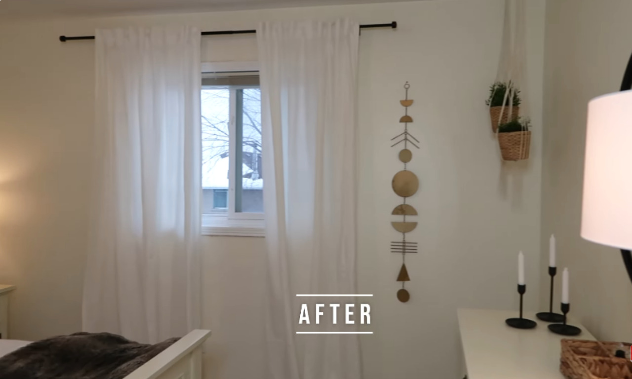 A view of a bedroom wall with a centered window covered by sheer white curtains hanging from a black rod. To the right of the window, a wall hanging featuring a vertical series of connected gold and wooden geometric shapes (half circles, lines, and full circles) is displayed. Below the wall hanging are two small woven baskets holding hanging plants. The word "AFTER" is overlaid in the bottom center.