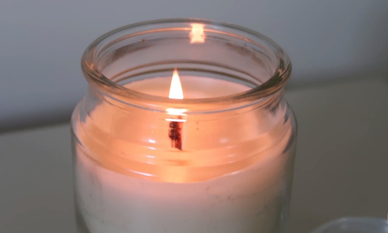 A close-up shot of a lit wick in a clear glass jar candle with white wax, casting a warm, soft glow.
