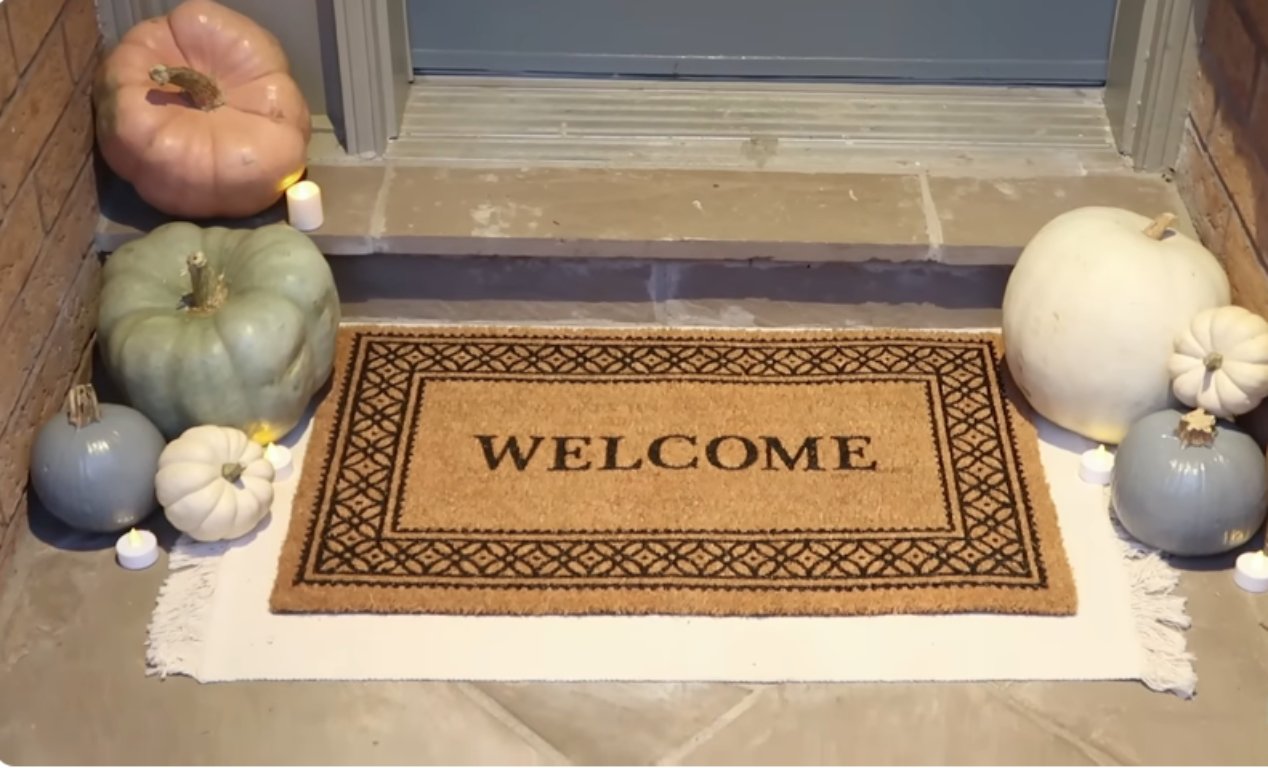 A close-up view of a front door threshold with a coir doormat that reads "WELCOME" centered on a light-colored rug. An arrangement of pastel pumpkins in various sizes and colors (pink, sage green, dusty blue, and white) flank the mat, with small white tea lights placed around them on the stone step.
