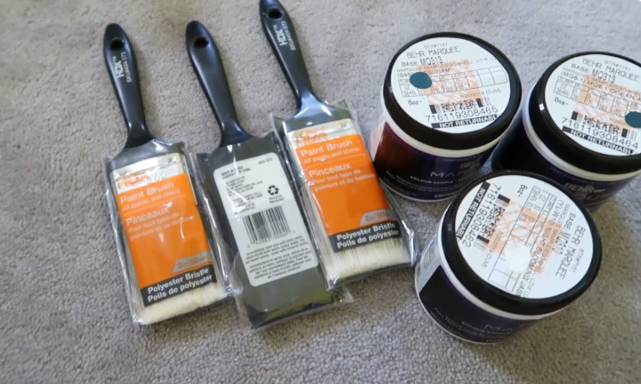 Three unopened paintbrushes with black handles and clear/orange packaging lying next to three small, round paint sample containers. The containers are labeled BEHR MARQUEE and have various color codes and stickers on them.
