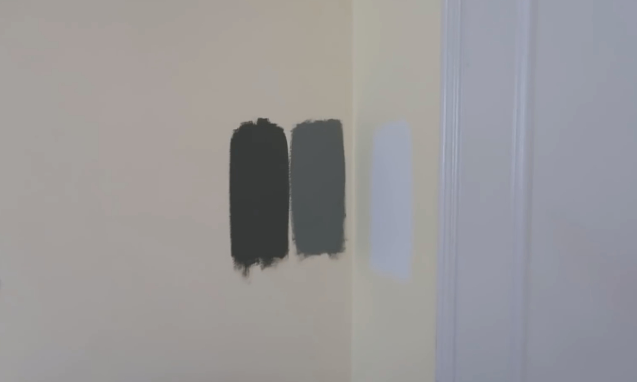 A close-up of a wall painted in a light beige or off-white color, displaying three vertical swatches of different paint colors brushed onto the surface. From left to right, the swatches are a very dark charcoal gray, a medium-dark gray, and a pale lavender or light blue-gray. A white door frame or trim is visible on the right side of the image.
