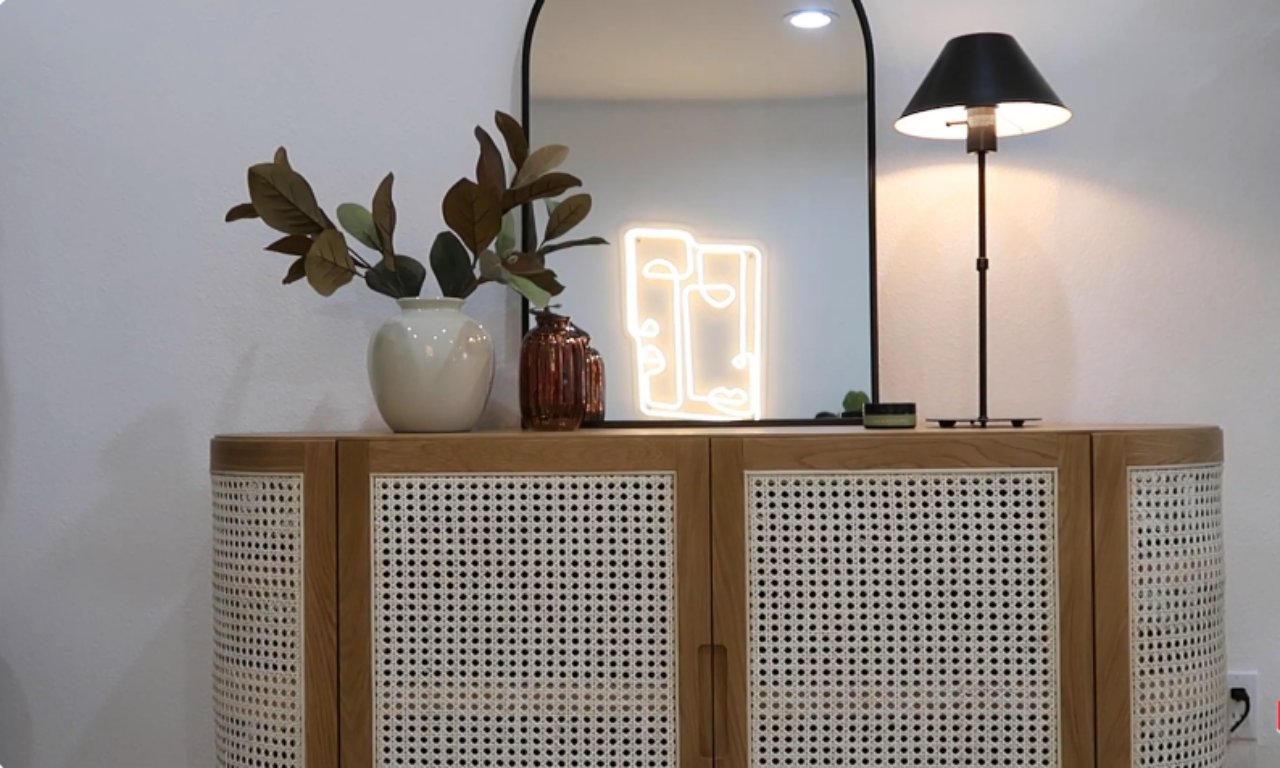 A detailed shot of a light wood and cane-fronted cabinet/console against a white wall. On top of the cabinet sits a tall black arched mirror, a white vase with decorative branches, a reddish-brown glass vase, a custom neon sign featuring abstract faces, and a modern black table lamp.