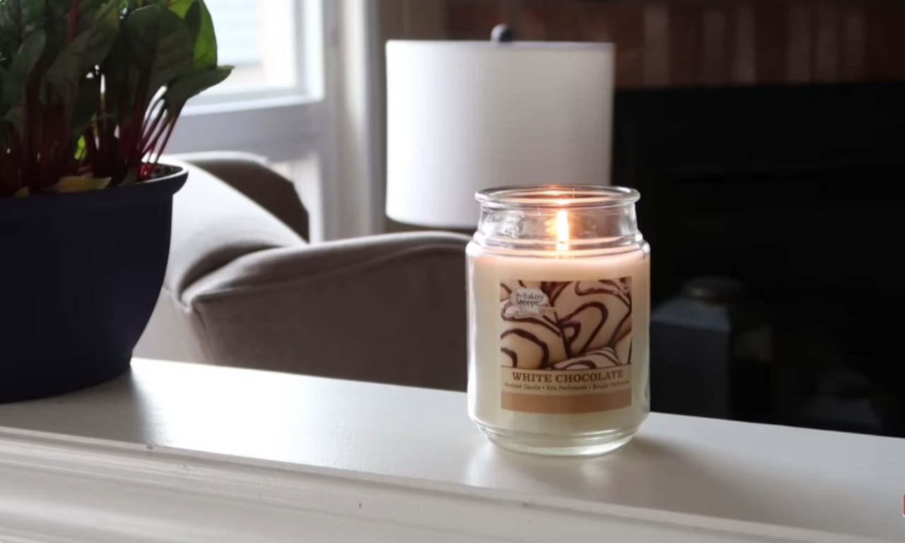 A lit candle in a clear glass jar labeled "WHITE CHOCOLATE" sitting on a white ledge or mantelpiece in a living room, with a potted plant and blurred furniture in the background.