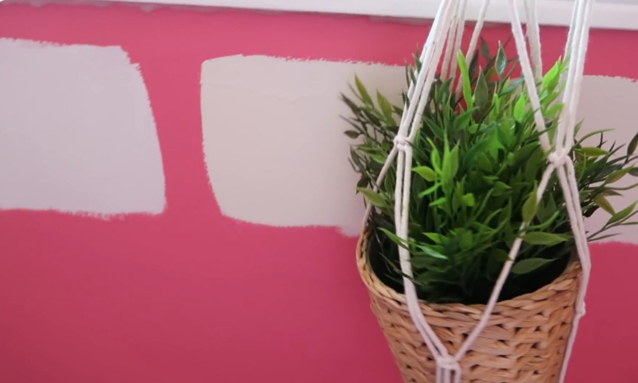 A close-up view of two horizontal swatches of a light, off-white paint color tested directly onto a bright magenta-pink wall. A hanging macrame planter with green faux greenery is in the foreground, partially obscuring the paint swatches, showing how decor would look against the tested colors.