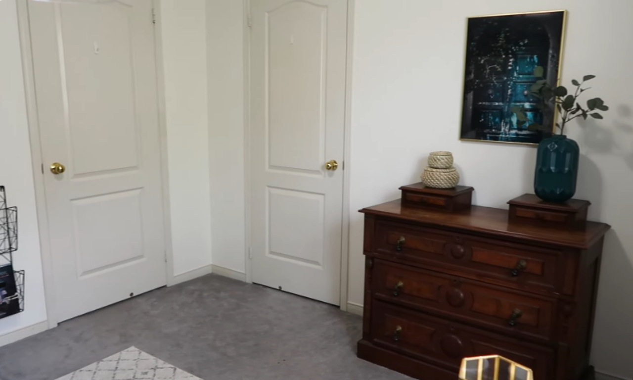 A dark wooden chest of drawers with three large drawers is placed against a white wall in the corner of a room, topped with two small woven baskets and a dark teal glass vase holding a leafy sprig. Two white doors with brass knobs are visible to the left. The room has light grey carpet.