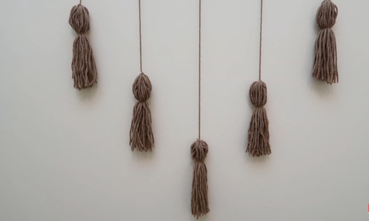 A close-up of a simple DIY tassel garland against a white wall. Five fluffy, brown yarn tassels of slightly different lengths hang vertically from individual strands of yarn. The tassels are gathered near the top, creating a distinct head and tail.