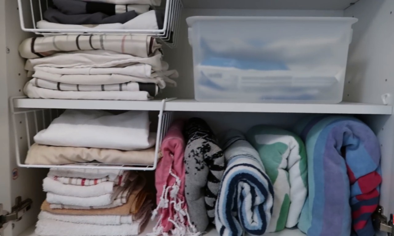  A white cabinet shelf used for linen storage. On the top shelf, a stack of folded sheets or towels sits next to a large, clear plastic storage bin. On the bottom shelf, items are stored on under-shelf wire baskets and directly on the shelf. The wire baskets hold folded neutral-colored sheets/linens. The bottom right section of the shelf holds several colorful, rolled-up towels (beach or bath towels).