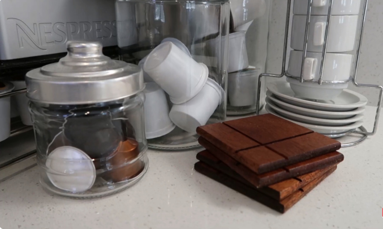 A close-up of a kitchen countertop featuring coffee accessories. On the left are two glass jars filled with assorted Nespresso-style coffee capsules. To the right is a stack of dark wooden coasters next to a chrome rack holding stacked white espresso cups and saucers.