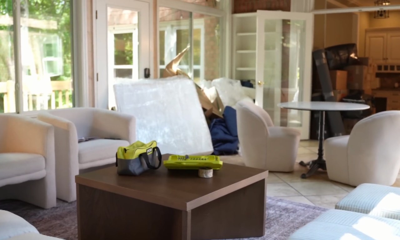 An indoor living space, likely a sunroom, filled with unpacked moving materials, boxes, and bubble wrap piled in the background. In the foreground, there are two white upholstered armchairs, a square dark brown wooden coffee table, and a small round black pedestal table. A green and black tool bag is resting on the coffee table.