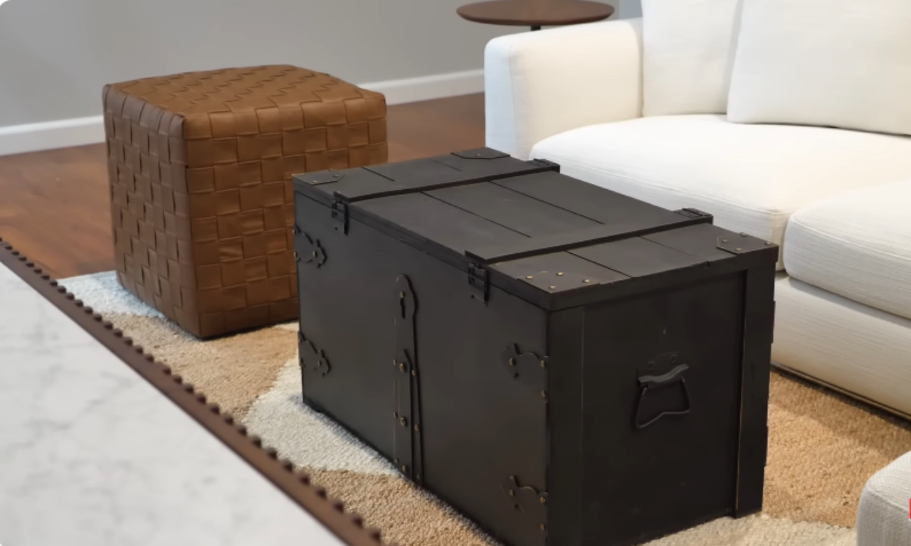 A close-up view of a living room scene featuring a dark wooden storage trunk used as a coffee table, a woven brown leather pouf, and a fluffy rug with beige, white, and brown tones layered over a wood floor. A white sectional sofa is partially visible in the background.